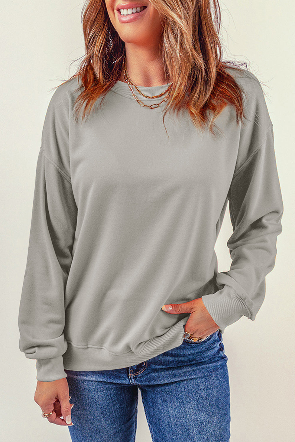 Person wearing a Gray Plain Crew Neck Pullover Sweatshirt and blue jeans with a hand in their pocket, adorned with layered necklaces.