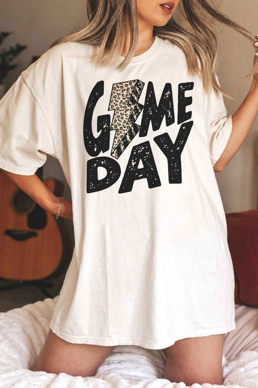 A person wearing a white "LEOPARD LIGHTNING GAME DAY Graphic Tee" with a leopard print lightning bolt design stands with a denim jacket partially on, showcasing its unisex sizing.