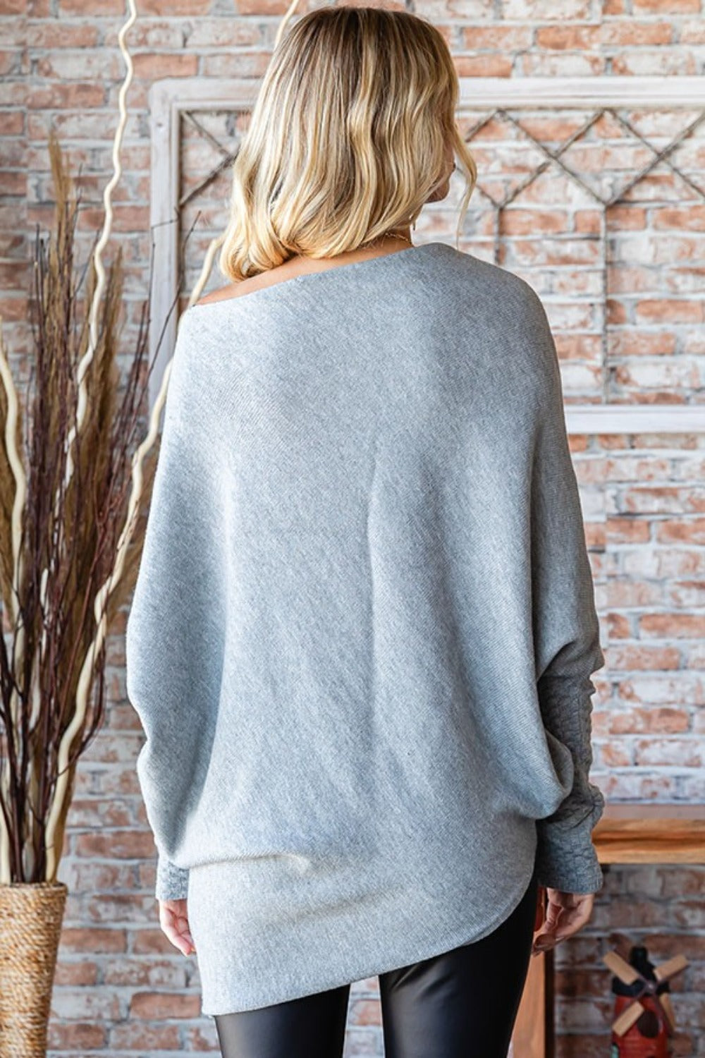 A woman wearing the First Love Full Size Asymmetrical Hem Dolman Sleeve Sweater, styled in a cozy and loose off-shoulder gray design, paired with black leggings, is standing indoors in front of a brick wall with decorative branches adorning the background.