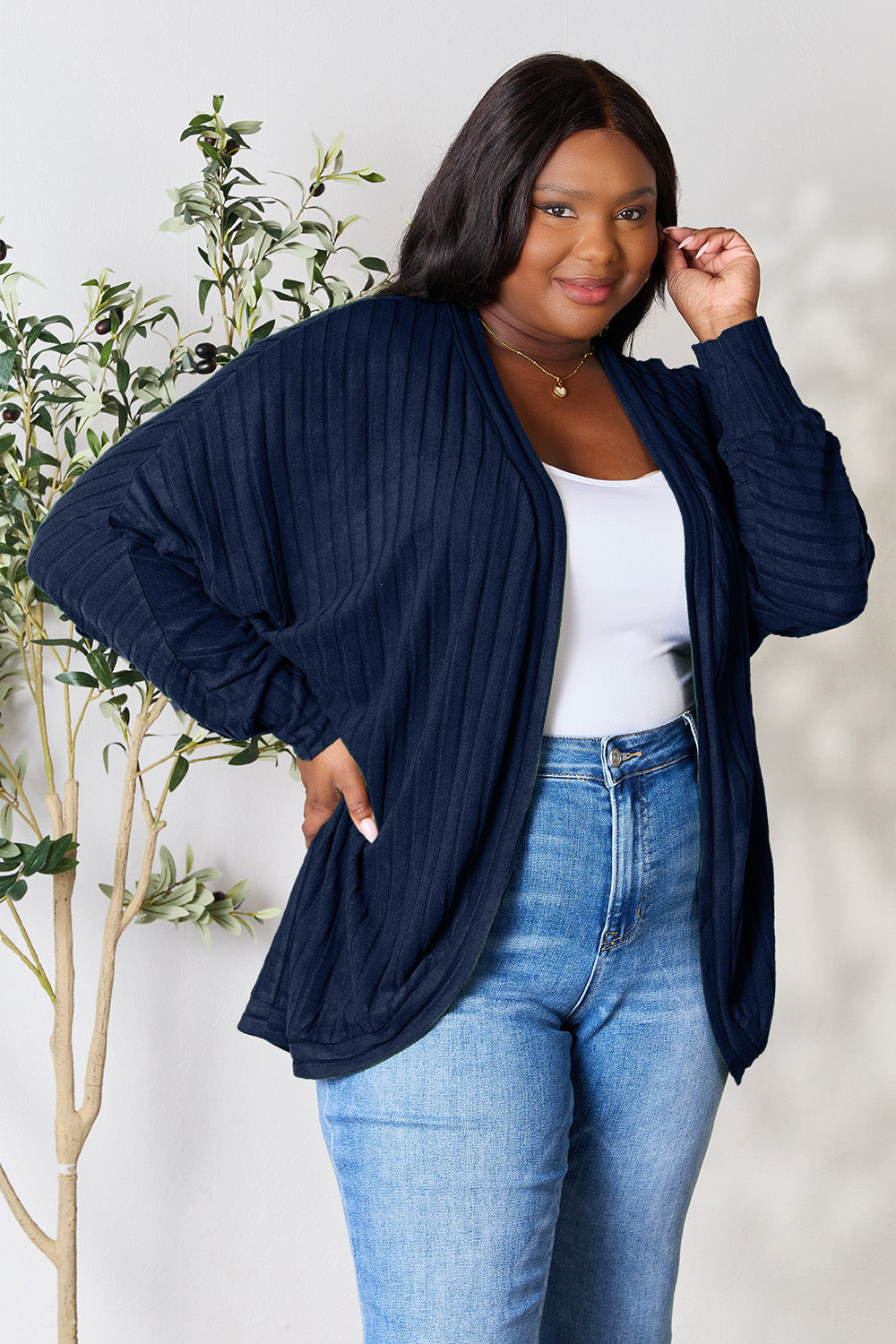 A person with wavy hair, wearing the Basic Bae Full Size Ribbed Cocoon Cardigan over a white top and beige pants, stands in front of a light-colored background with a leafy plant, showcasing an effortlessly basic style.