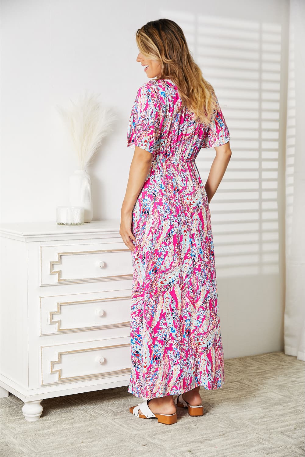 A woman stands in a room wearing the Double Take Multicolored V-Neck Maxi Dress, paired with white sandals. A dresser and curtain create a charming backdrop.