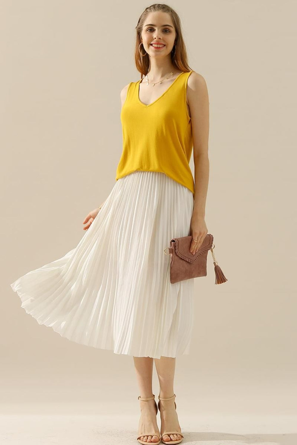 A woman in a stylish and versatile Ninexis Full Size V-Neck Curved Hem Tank and white pleated skirt poses while holding her handbag.