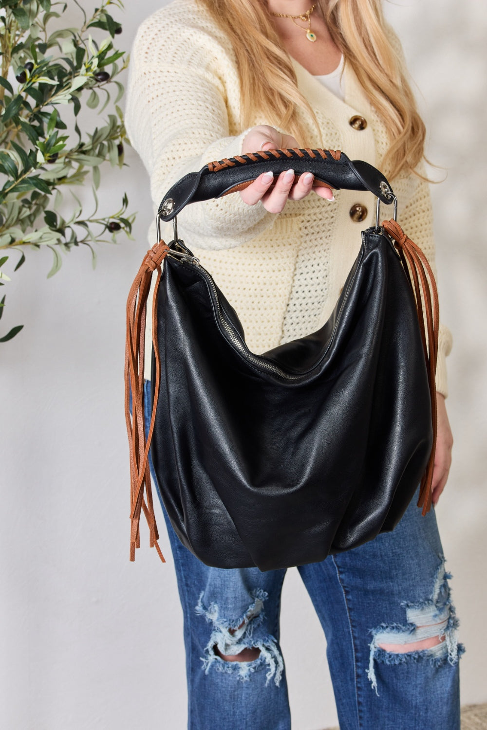 Individual holding a SHOMICO Fringe Detail Contrast Handbag, featuring cream and brown accents, paired with a knitted cream sweater, standing against a light background. The handbag exudes Bohemian flair with its relaxed style.