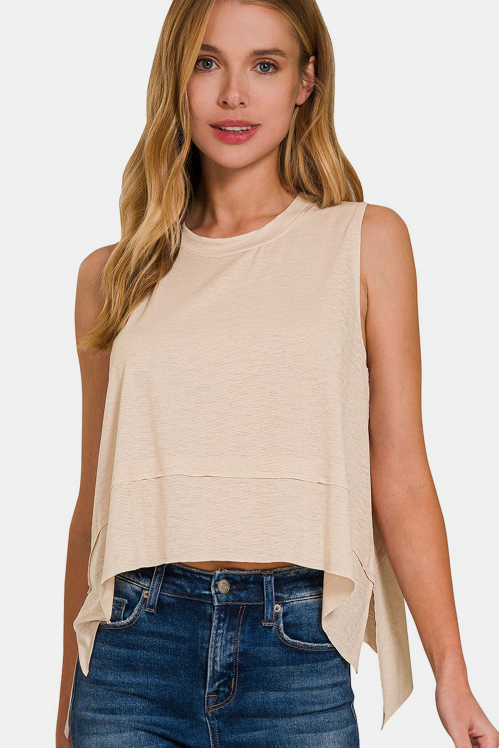 A woman with long, light brown hair wears a versatile piece: the Zenana Exposed Seam Slit Round Neck Tank in beige paired with blue jeans, standing against a plain background.