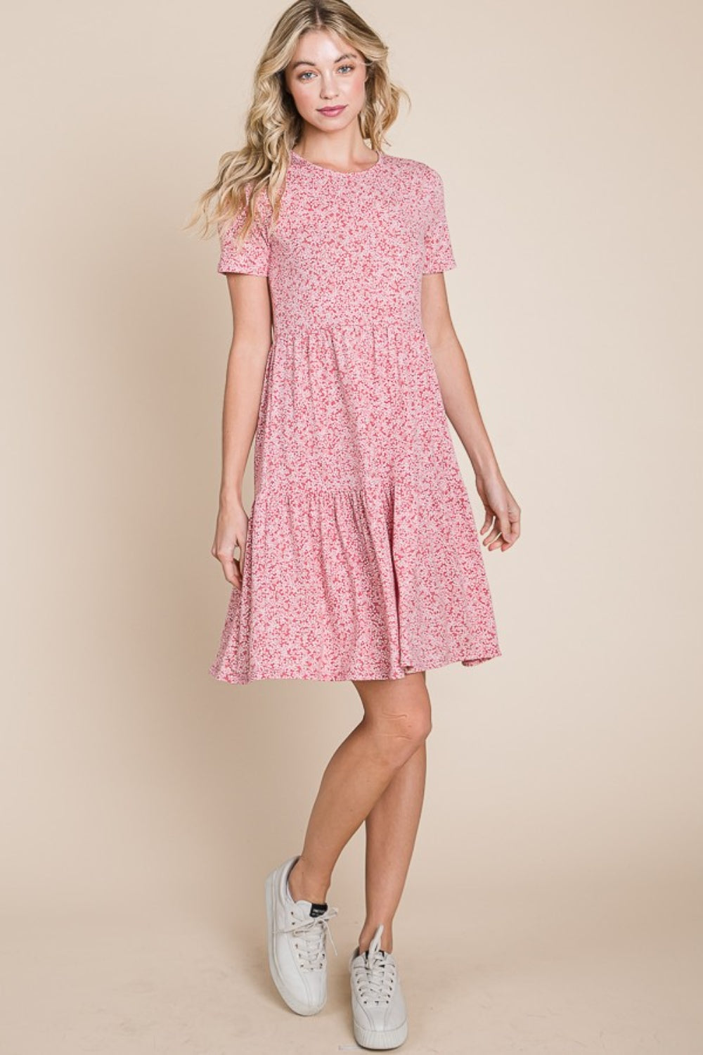 A woman with wavy blonde hair is smiling and holding the side of her BOMBOM Printed Short Sleeve Mini Dress, a lightweight summer dress featuring a pink pattern.