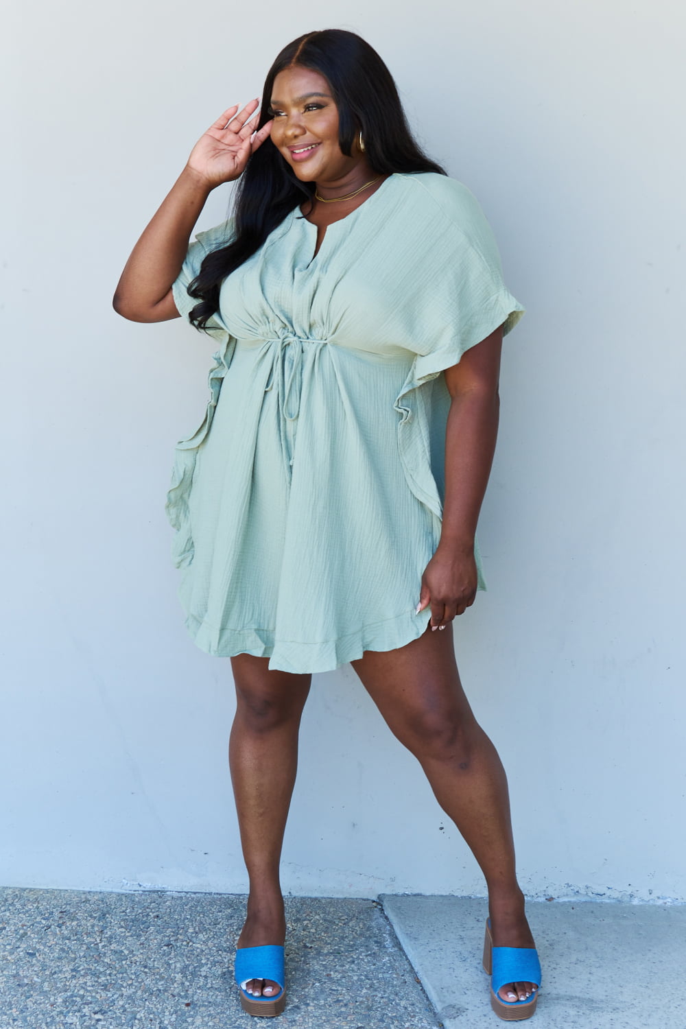 A woman is wearing the Ninexis Out Of Time Full Size Ruffle Hem Dress with Drawstring Waistband in Light Sage. Her long, dark hair falls gracefully as she smiles and looks to her left, standing against a plain background.