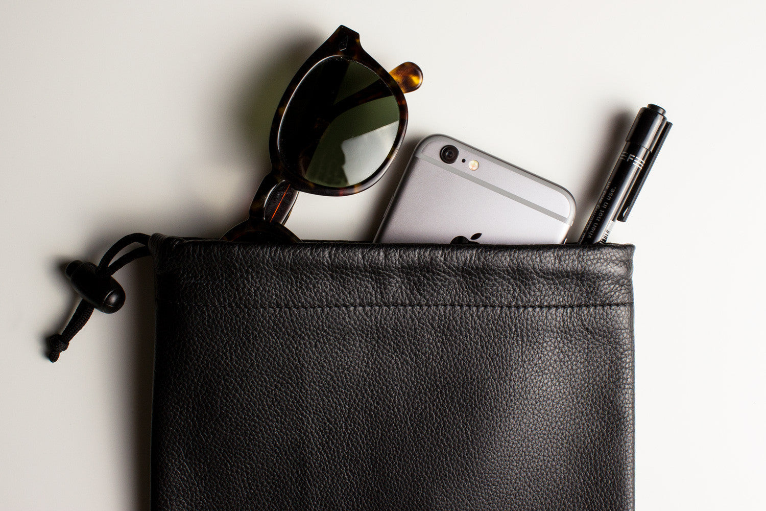 A sophisticated black Accessory Pouch, fashioned from high-quality cowhide leather, partially open to display a smartphone, a chocolate bar, and an additional small item inside. The adjustable closure provides a secure fit for all your essentials.