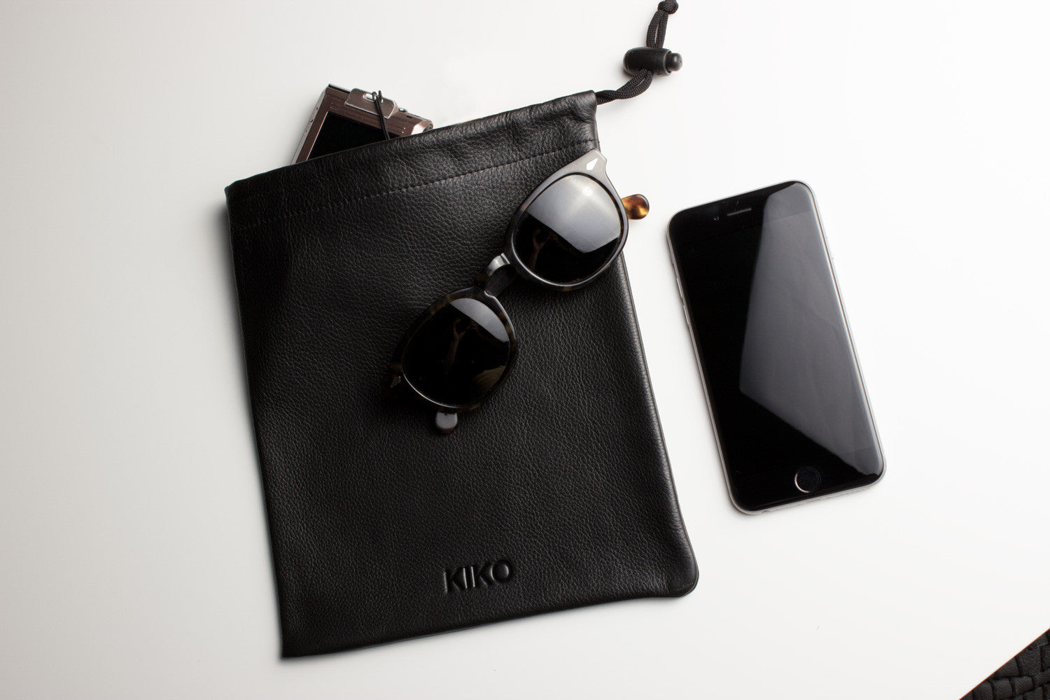 A sophisticated black Accessory Pouch, fashioned from high-quality cowhide leather, partially open to display a smartphone, a chocolate bar, and an additional small item inside. The adjustable closure provides a secure fit for all your essentials.