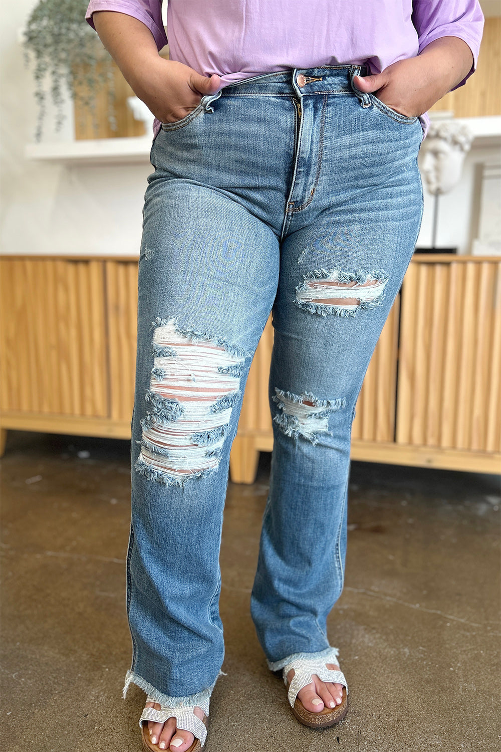 A person wearing Judy Blue Full Size Distressed Raw Hem Bootcut Jeans and a light purple top stands on a polished floor with a wooden cabinet in the background.