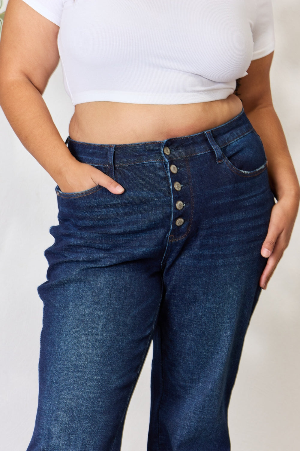 A person stands with hands in pockets, wearing the Judy Blue Full Size Button-Fly Straight Jeans, known for their high-waisted dark blue design crafted from premium lycra fabric. The jeans showcase button details and a straight leg cut, complemented by a white top and open-toed heels.