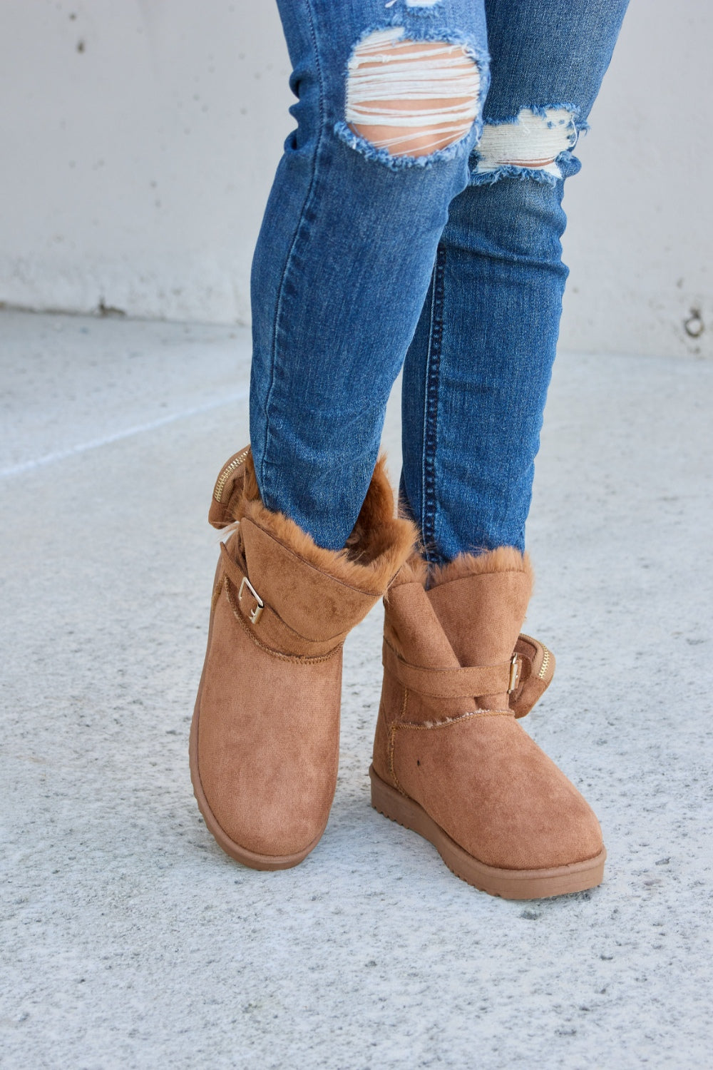 Person wearing ripped blue jeans and Forever Link Plush Thermal Flat Boots, tan in color, fur-lined with side buckles, standing on a concrete surface. These stylish boots promise cozy insulation for the feet.