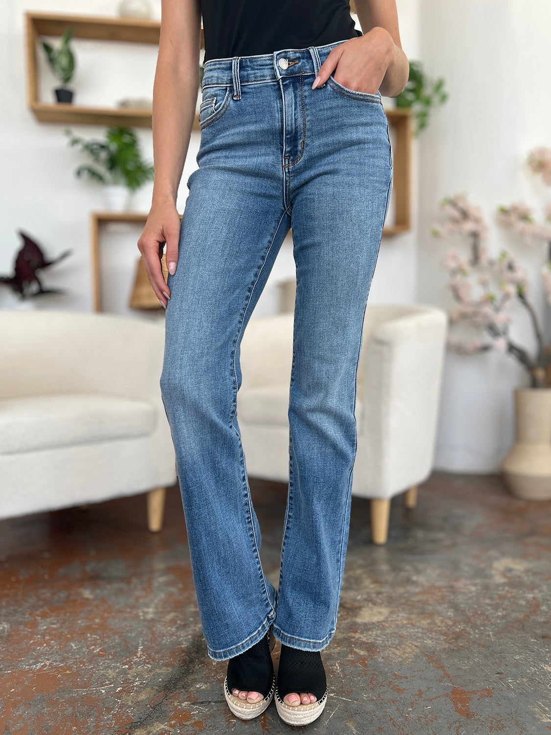 Person wearing Judy Blue Full Size Mid-Rise Waist Straight Jeans and a black top, standing in a room with white chairs, potted plants, and wooden shelves. Only the lower half of the body and part of the arms are visible.