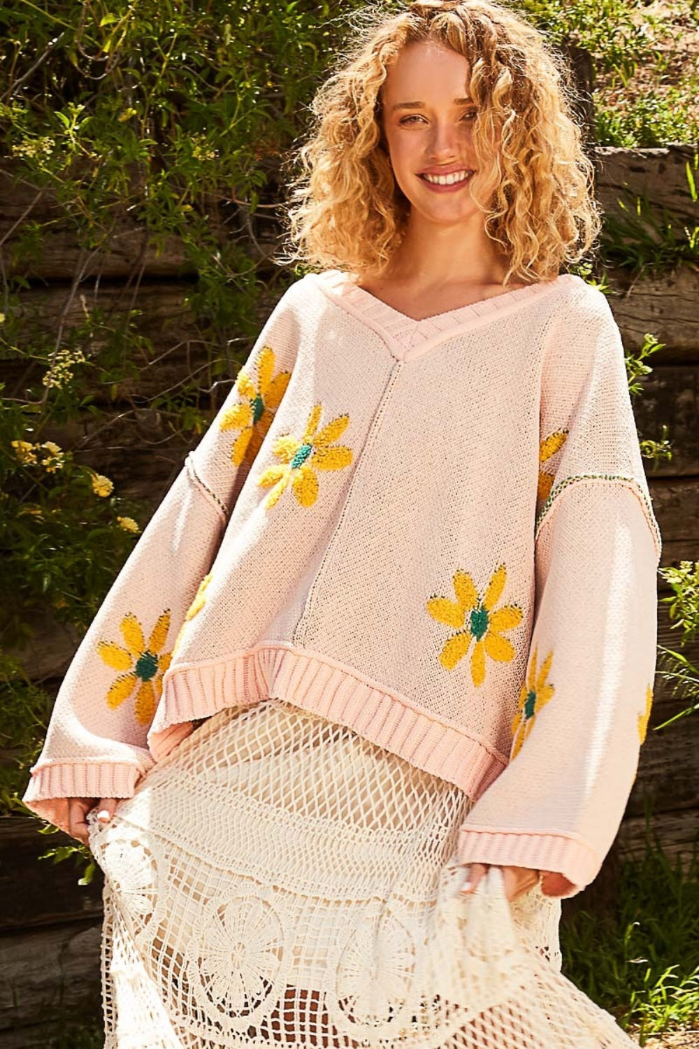 A woman with curly hair smiles while wearing a cozy POL V-Neck Floral Pattern Chenille Sweater in pink, adorned with yellow flower patterns, and holding a netted skirt in an outdoor setting.