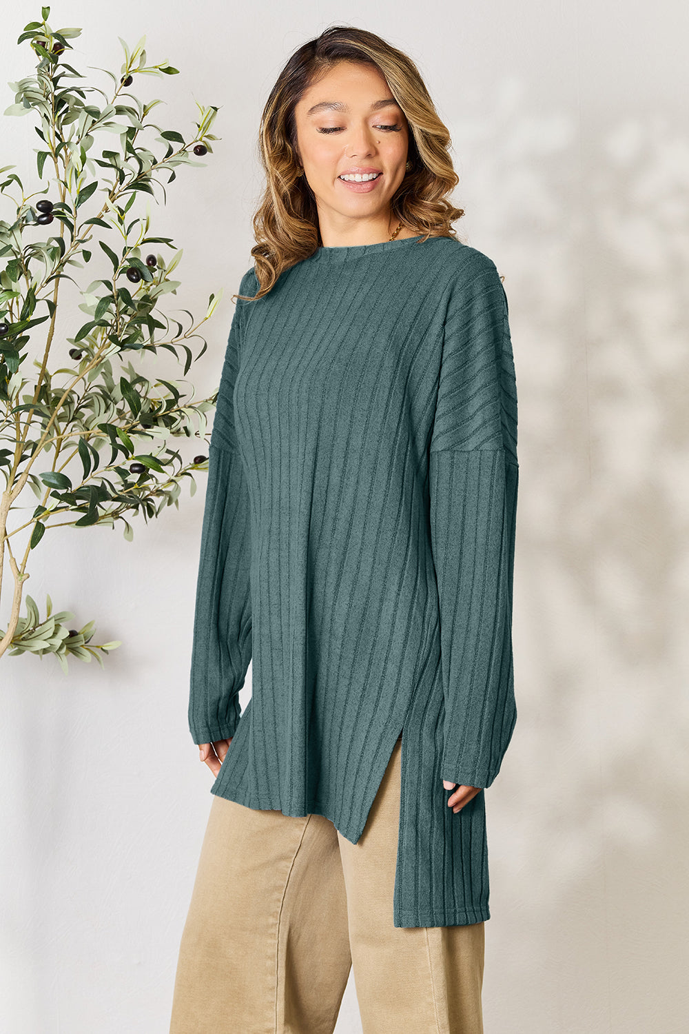 A person with shoulder-length hair smiles while wearing a beige ribbed sweater called the Basic Bae Full Size Ribbed Round Neck Long Sleeve Slit Top, along with matching beige pants, showcasing a basic style. They stand next to a green plant.