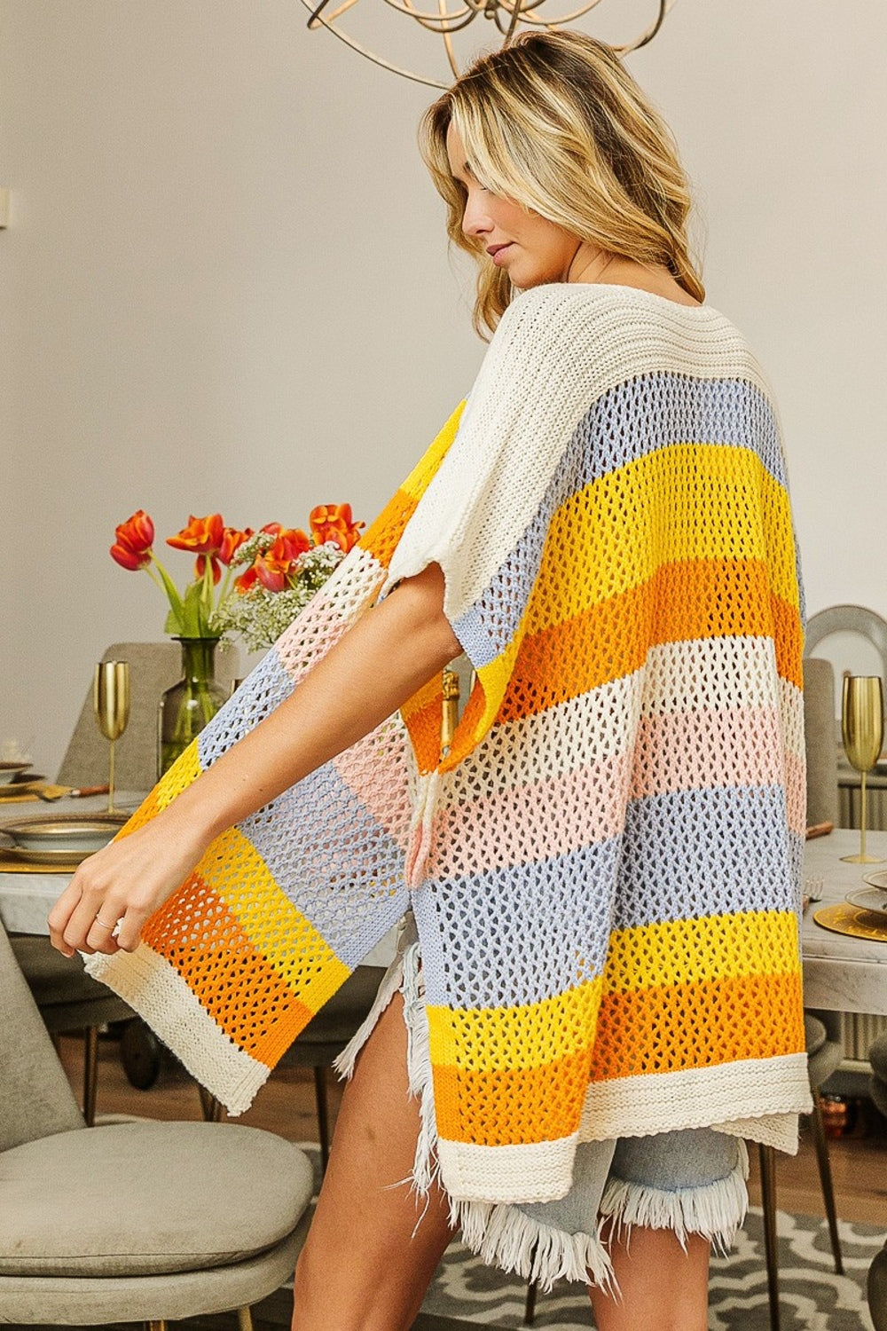 A woman in a brightly colored BiBi Openwork Striped Open Front Knit Cardigan and shorts stands in a dining room with a set table and a vase of flowers, showcasing this versatile layering piece.