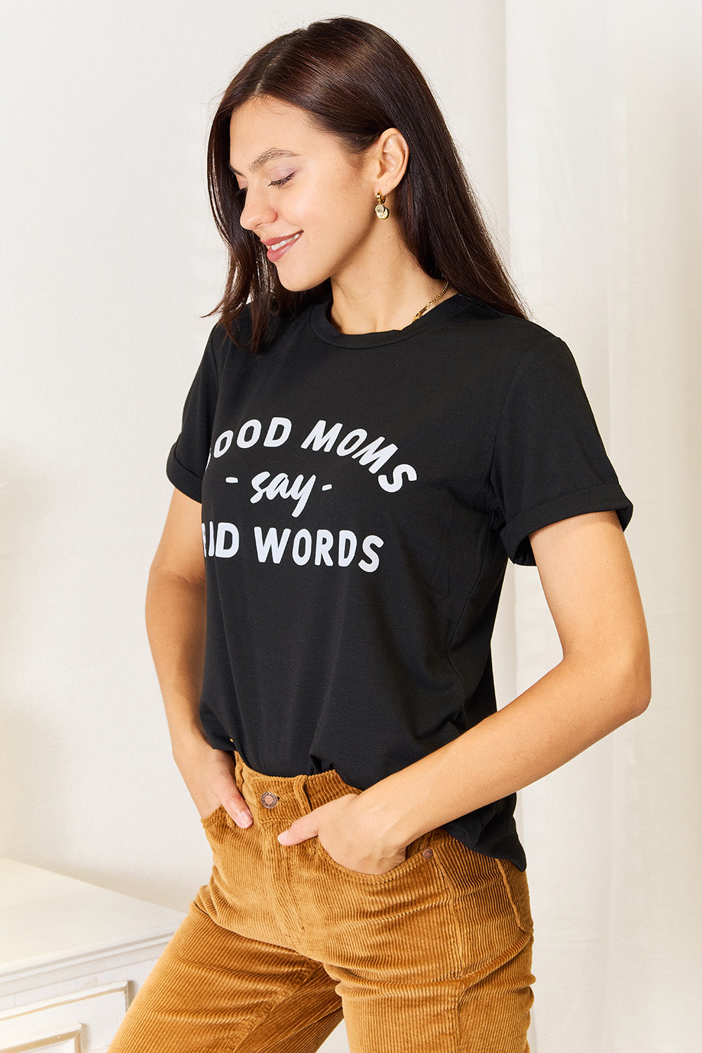 A woman stands in a room with a white wall, where light filters through the blinds, subtly showcasing her rebellious side while wearing the Simply Love GOOD MOMS SAY BAD WORDS Graphic Tee.