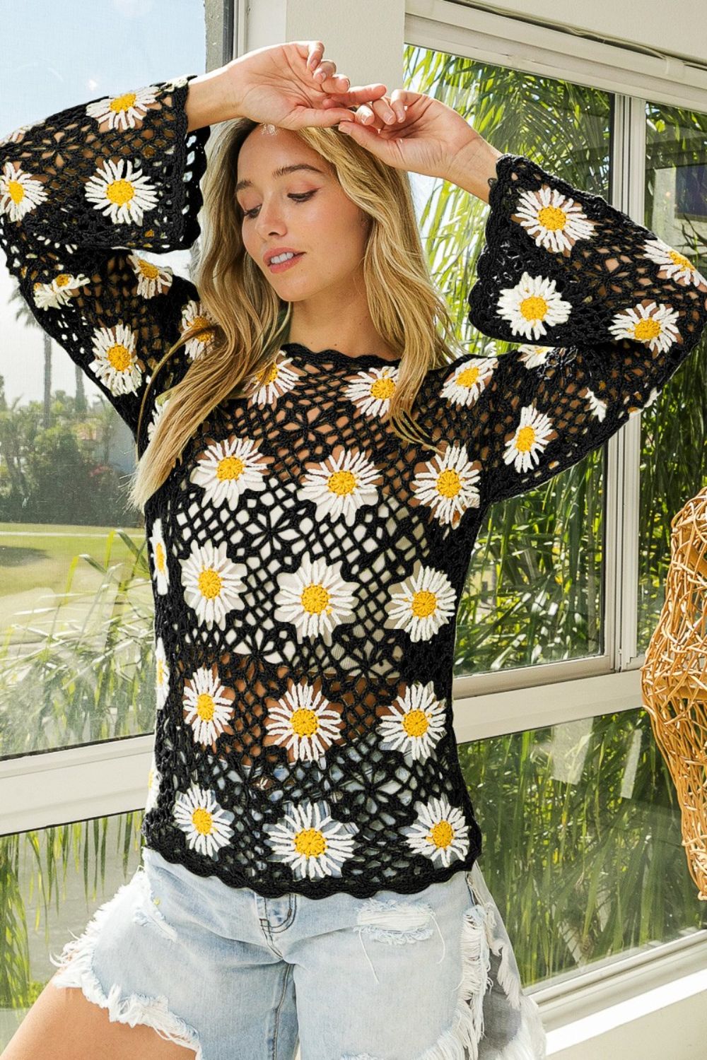 A person poses near a window wearing the BiBi Floral Crochet Net Lace Cover Up with white and yellow flower patterns and light blue distressed denim shorts—a perfect addition to summer outfits.