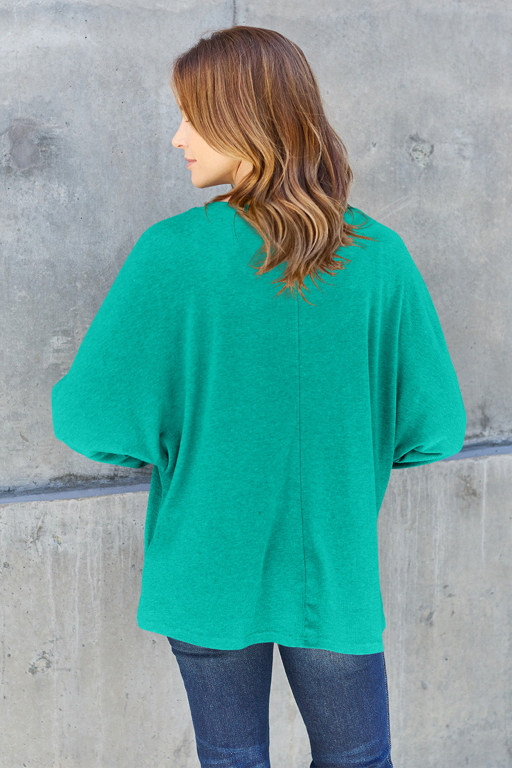 A person wearing a Double Take Full Size Round Neck Long Sleeve T-Shirt in green, blue distressed jeans with a slightly stretchy fit, and a crossbody bag, holding a beverage in a plastic cup.