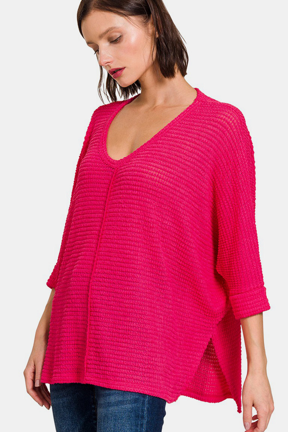 A woman wearing the Zenana V-Neck High-Low Jacquard Knit Top in a bright pink, loose-knit style with three-quarter sleeves, paired with blue jeans, stands against a plain white background, showcasing her comfortable knit top.