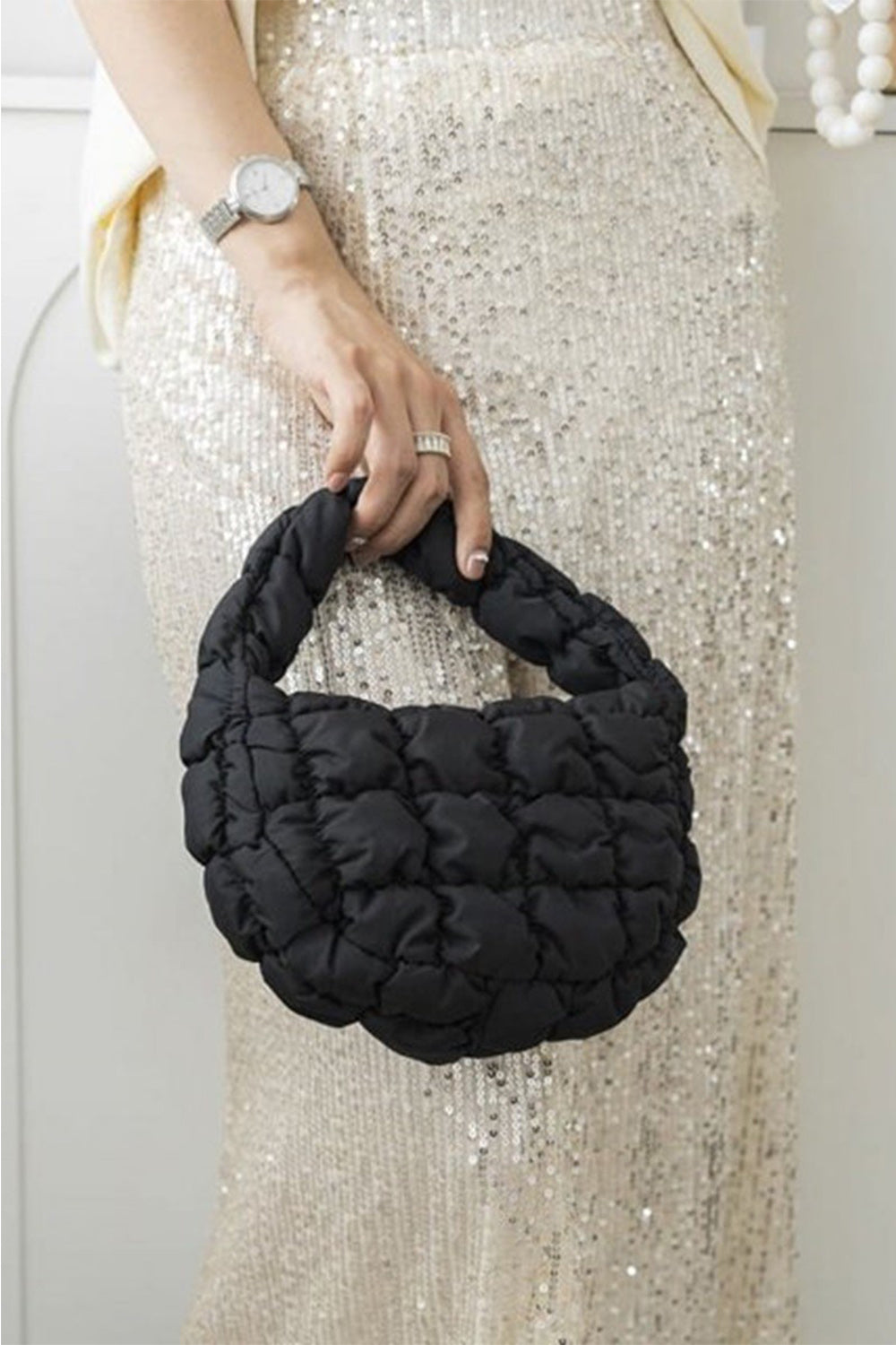 A person wearing a sequined dress holds the Zenana Quilted Micro Puffy Handbag with a ring handle, adding a stylish accessory to their look.