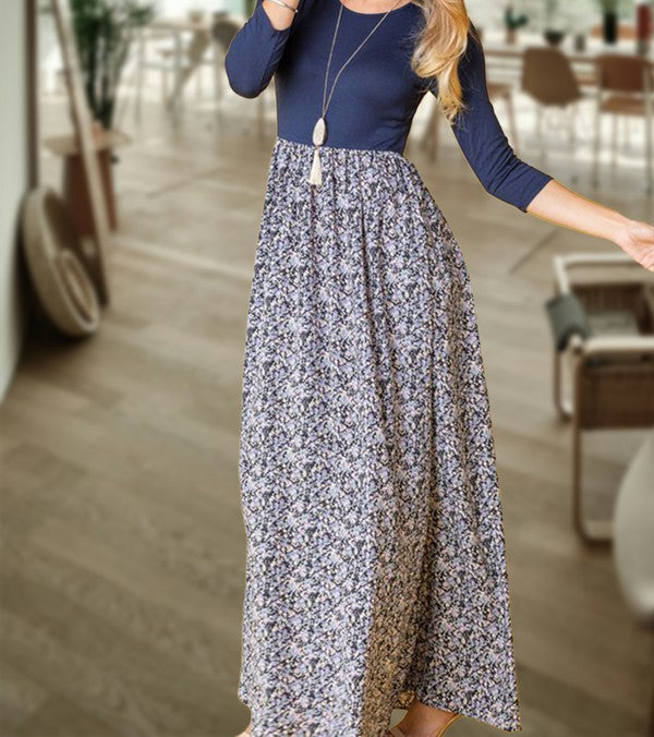 A woman elegantly stands in a room with wooden flooring and modern furniture, wearing a ditsy floral maxi dress and a long-sleeve navy top.