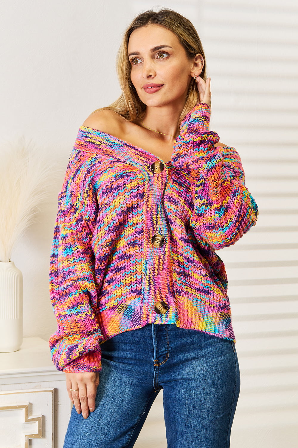 Person wearing an Angel Wings Woven Right V-Neck Long Sleeve Cardigan, characterized by its multicolored design and large buttons, paired with blue jeans. The cardigan, made of 100% acrylic knit, offers a versatile look perfect for any occasion. They are standing indoors against a white background.