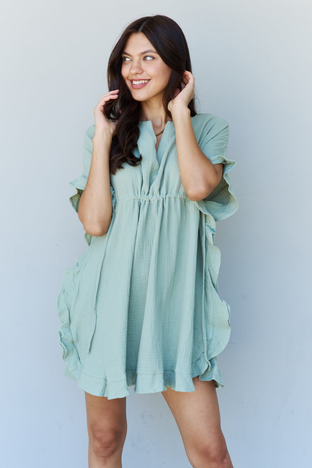 A woman is wearing the Ninexis Out Of Time Full Size Ruffle Hem Dress with Drawstring Waistband in Light Sage. Her long, dark hair falls gracefully as she smiles and looks to her left, standing against a plain background.