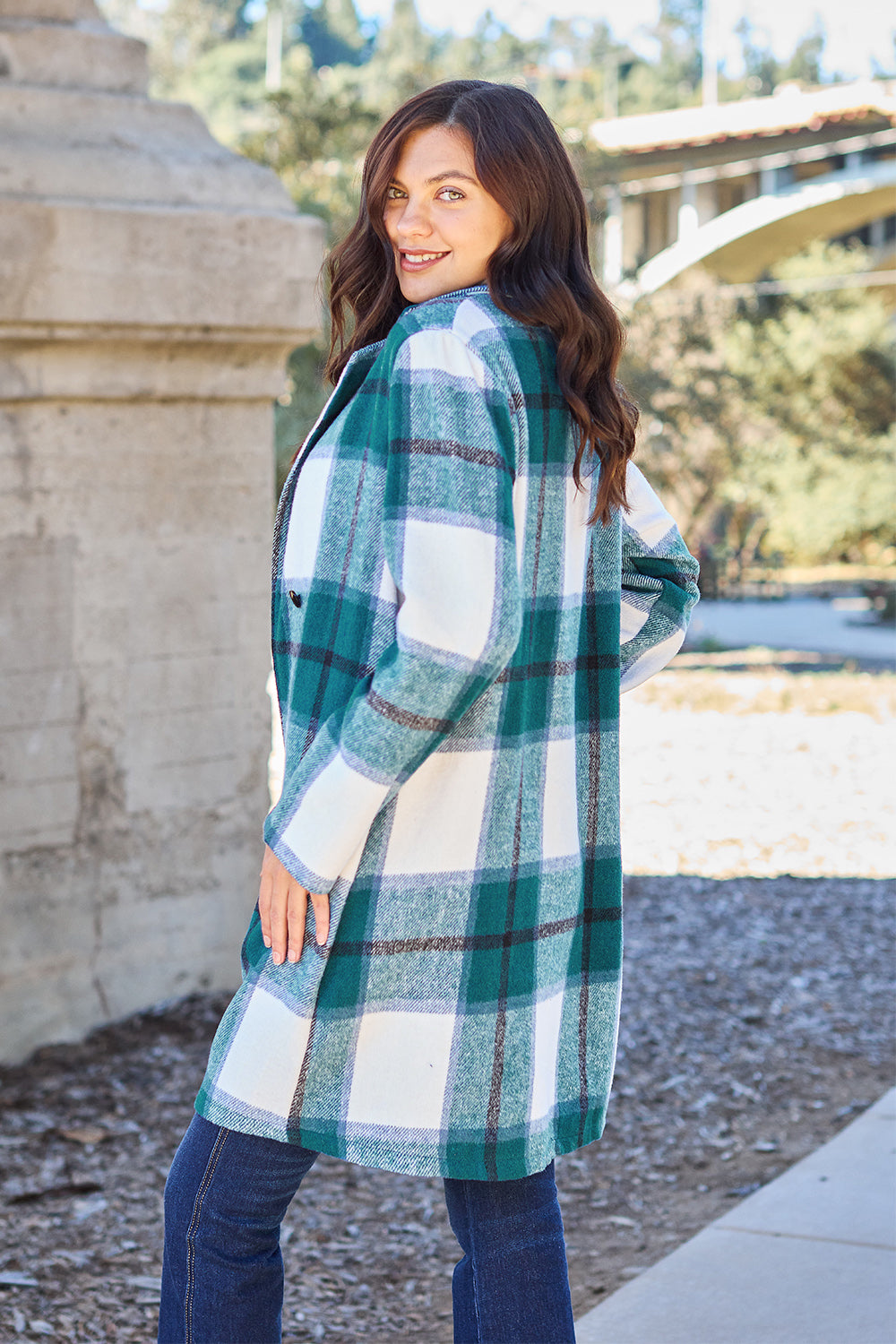 A woman stands on a sidewalk wearing the Double Take Full Size Plaid Button Up Lapel Collar Coat in pink and white over a white top and blue jeans, with her right hand lightly touching her hair. The polyester fabric of the coat makes it durable and perfect for an easy machine wash cold.