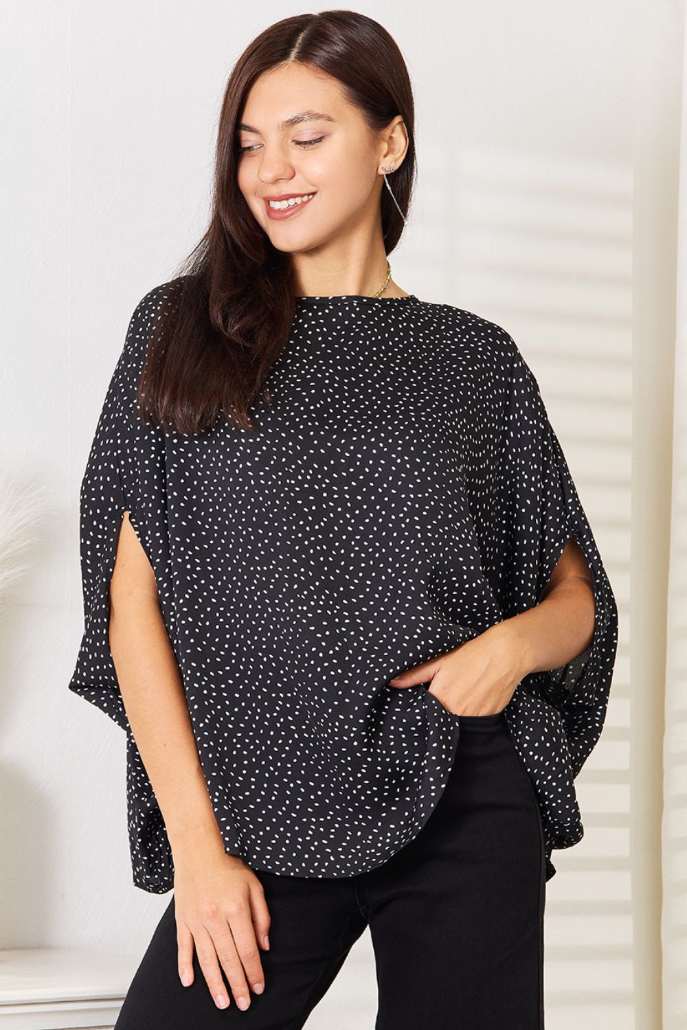 A woman stands wearing the Perfee Printed Dolman Sleeve Round Neck Blouse with white dots and black pants, smiling and looking to her left. The blouse features a printed design that adds a touch of elegance to her outfit.