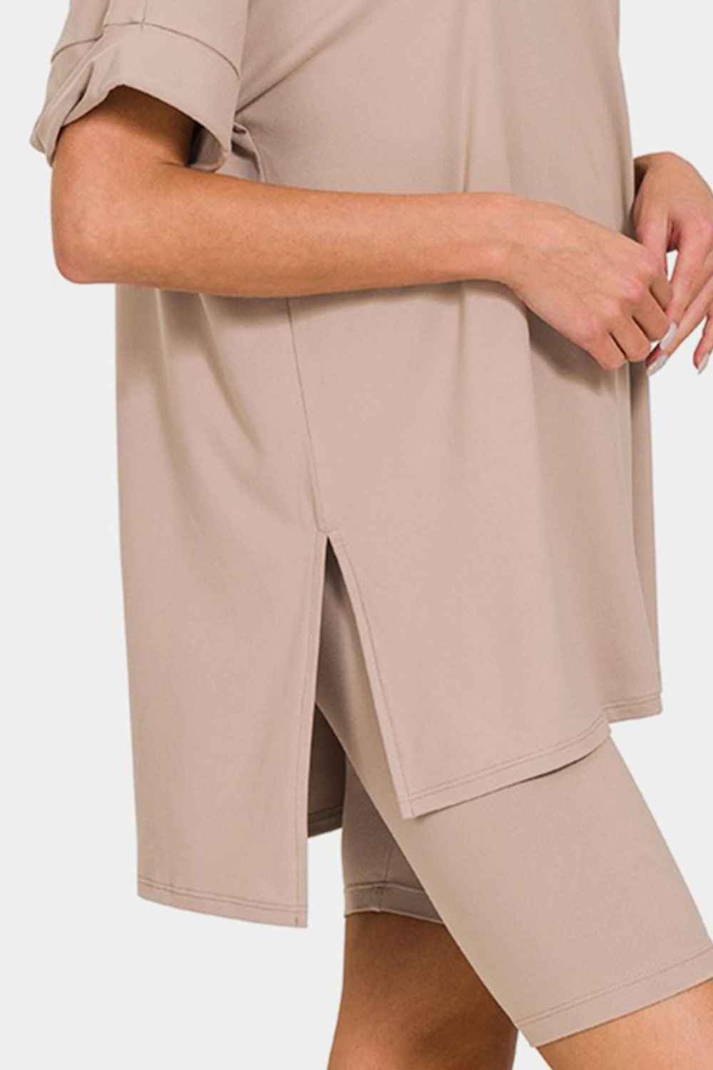 A woman wearing the highly stretchy Zenana Full Size V-Neck Short Sleeve Slit T-Shirt and Shorts Set in beige, featuring a mid-thigh shorts and short sleeve t-shirt combination, stands against a plain background.