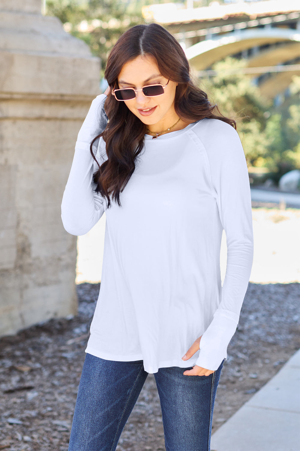 A woman wearing sunglasses and a slightly stretchy Basic Bae Full Size Round Neck Long Sleeve T-Shirt in white stands on a sidewalk with an arched structure in the background, smiling slightly.