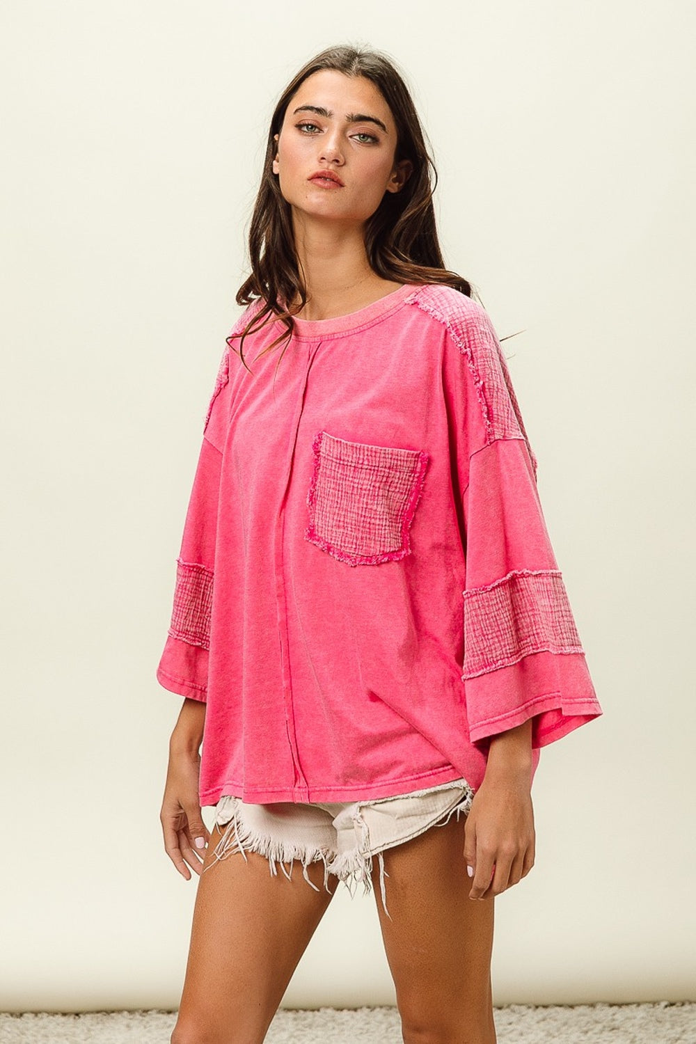 A young woman with long dark hair wears a bright pink BiBi High-Low Washed T-Shirt featuring a front pocket, paired with white frayed denim shorts. She poses against a plain background, effortlessly combining comfort and style in her trendy wardrobe.