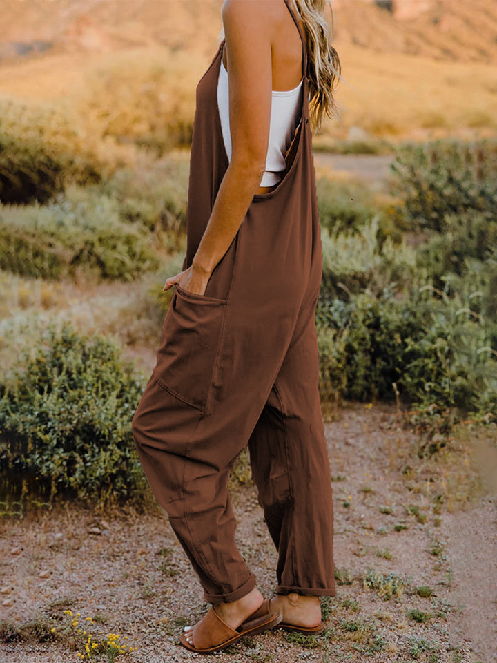 A woman stands outdoors wearing the Double Take Full Size Sleeveless V-Neck Pocketed Jumpsuit in a loose-fitting, imported brown design over a white top. Her surroundings include grass, bushes, and a rocky landscape in the background. The jumpsuit's polyester fabric ensures comfort and durability with every machine wash.