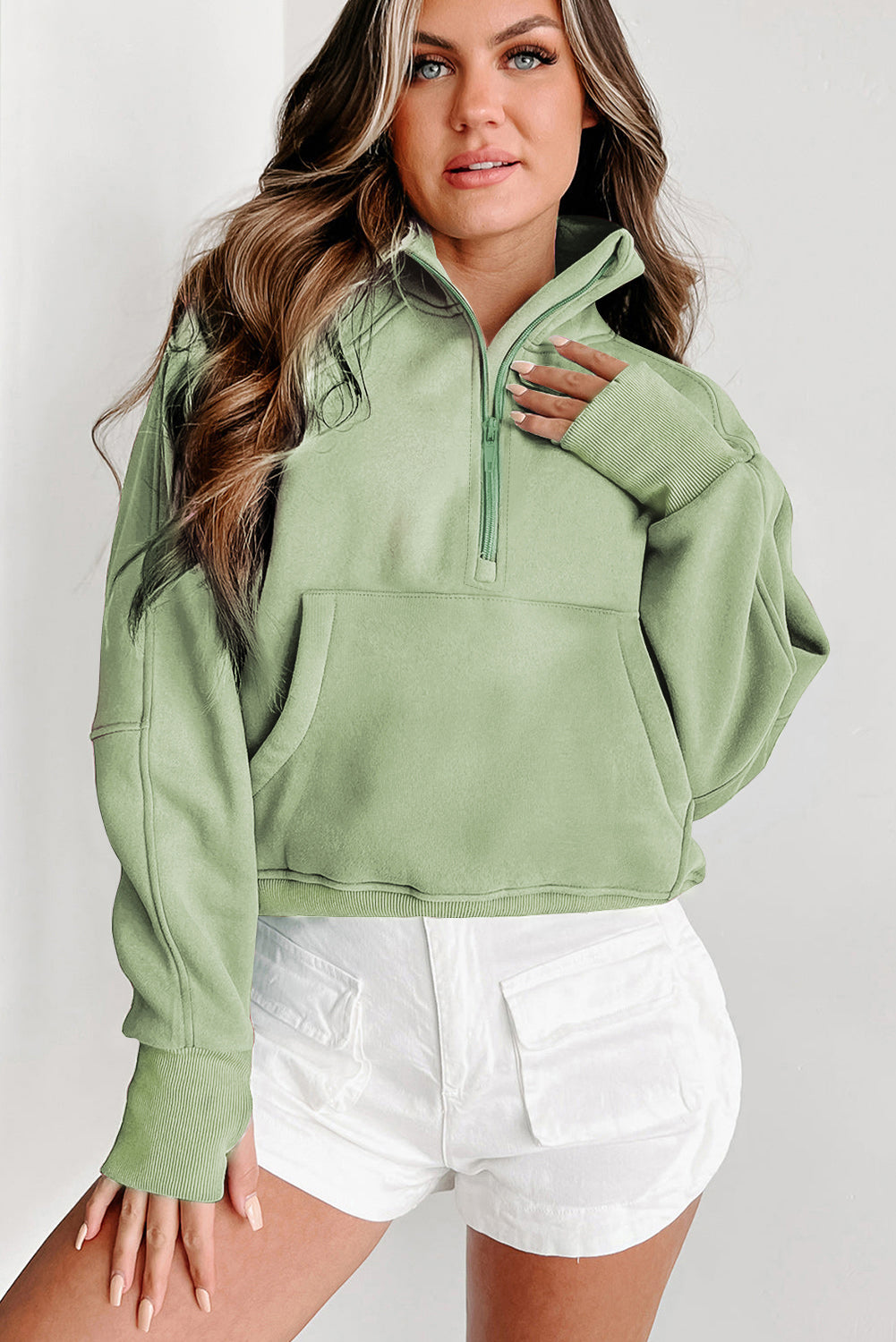 A woman with long wavy hair is wearing a chic Smoke Green Fleece Lined Zip Up Stand Collar Thumbhole Sleeve Sweatshirt and white shorts, facing sideways.