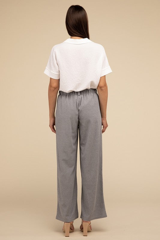 A woman stands against a beige background, highlighting her chic ensemble with a tailored fit. She wears a white short-sleeve shirt paired with versatile pink Waffle Trouser Pants, hands casually in pockets, complemented by stylish strappy sandals.