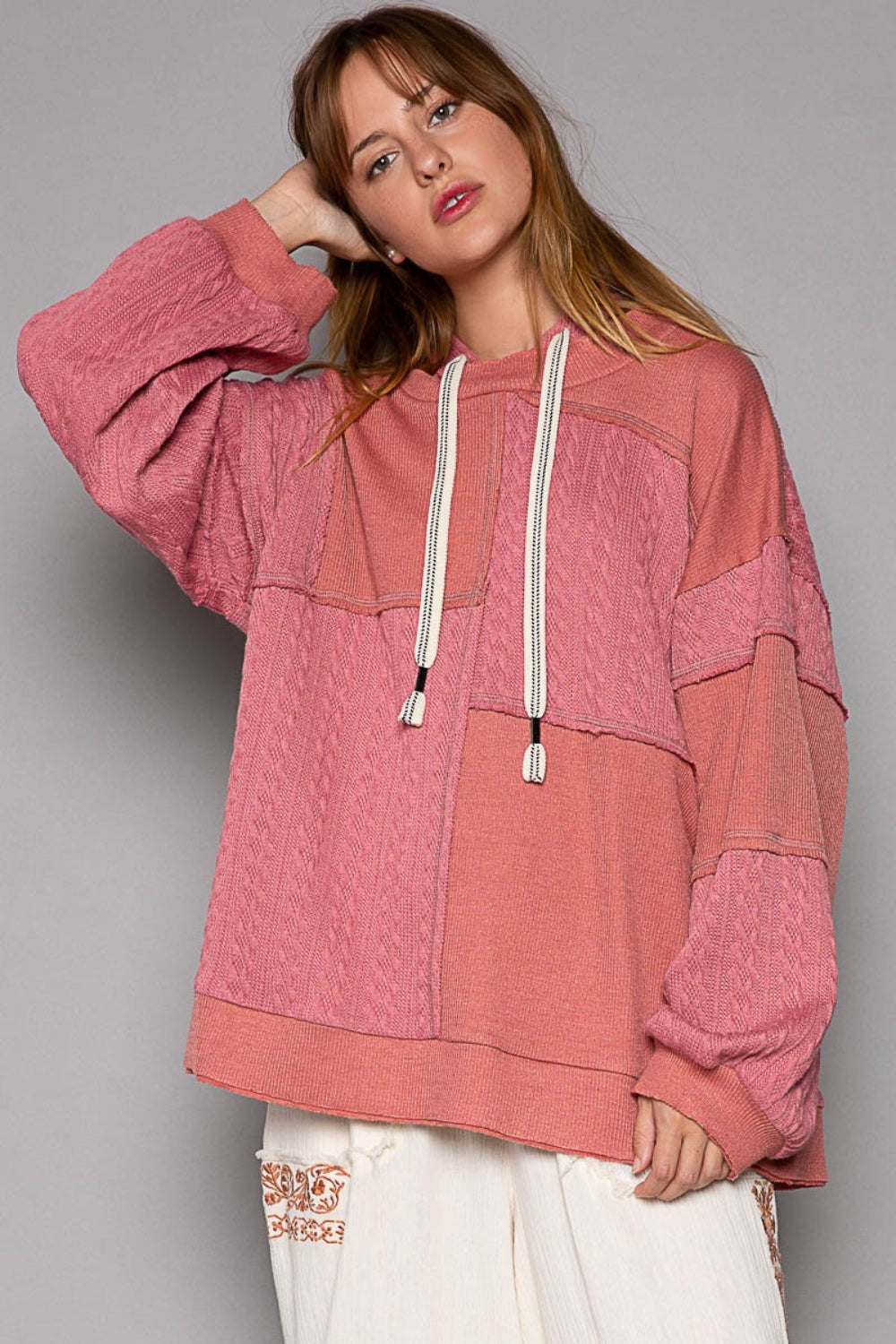 A person wearing the POL Exposed Seam Hooded Knit Top, featuring a pink patchwork design with balloon sleeves and white drawstrings, paired with relaxed-fit white pants, poses against a gray background.