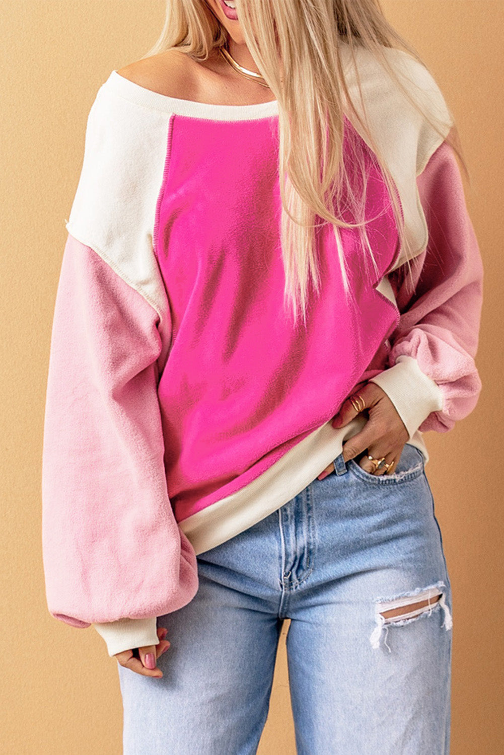 A person with long blonde hair is standing facing away from the camera against a beige background, showcasing the comfortable fleece fabric of the Rose Colorblock Long Sleeve Pullover Fleece Sweatshirt in pink shades and light blue jeans.