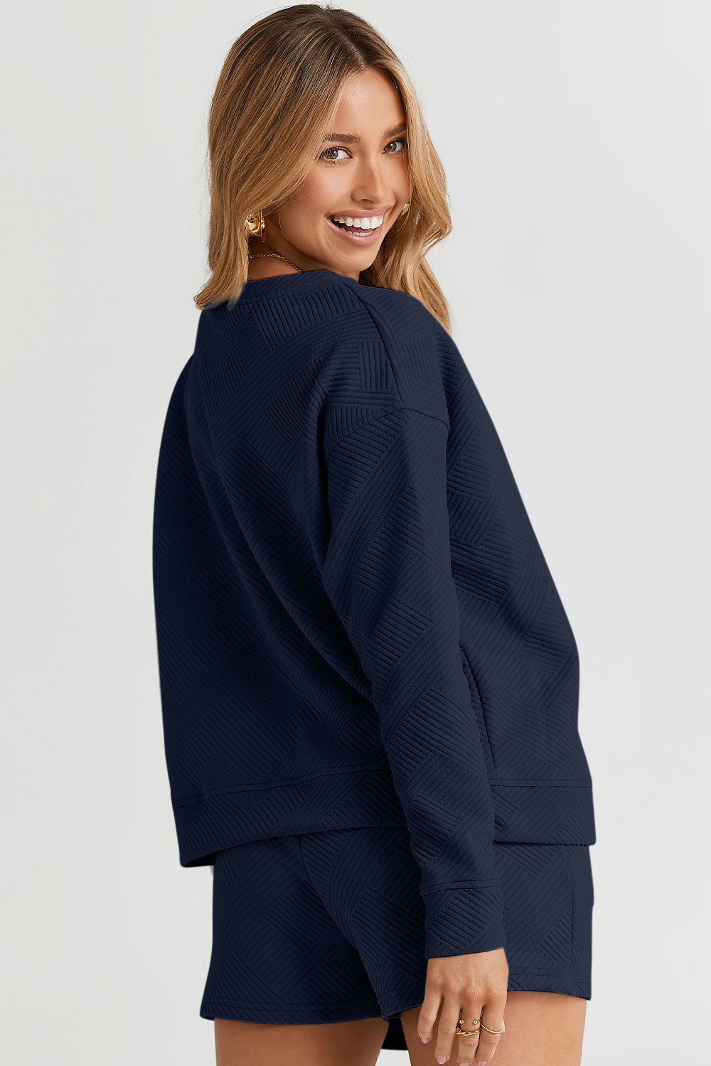 A woman decked out in the Double Take Full Size Texture Long Sleeve Top and Drawstring Shorts Set, featuring a brown textured two-piece with a long-sleeve top and matching pocketed shorts, smiles at the camera.