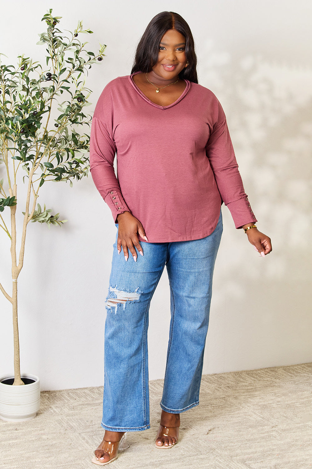 A person stands in front of a plain backdrop with a plant, wearing a maroon Culture Code Full Size V-Neck Exposed Seam Long Sleeve Blouse and blue jeans, smiling slightly with their hand on their hip. The relaxed fit of the polyester blend adds to the casual yet stylish look.