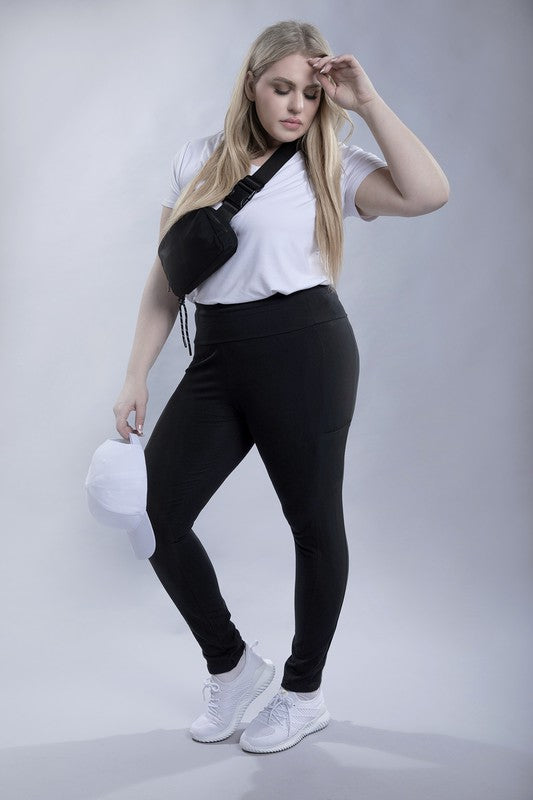 A person with long hair in casual attire is wearing a white t-shirt, Plus Everyday Leggings with Pockets in black, and white sneakers. They are holding a white cap and sporting a black crossbody bag.