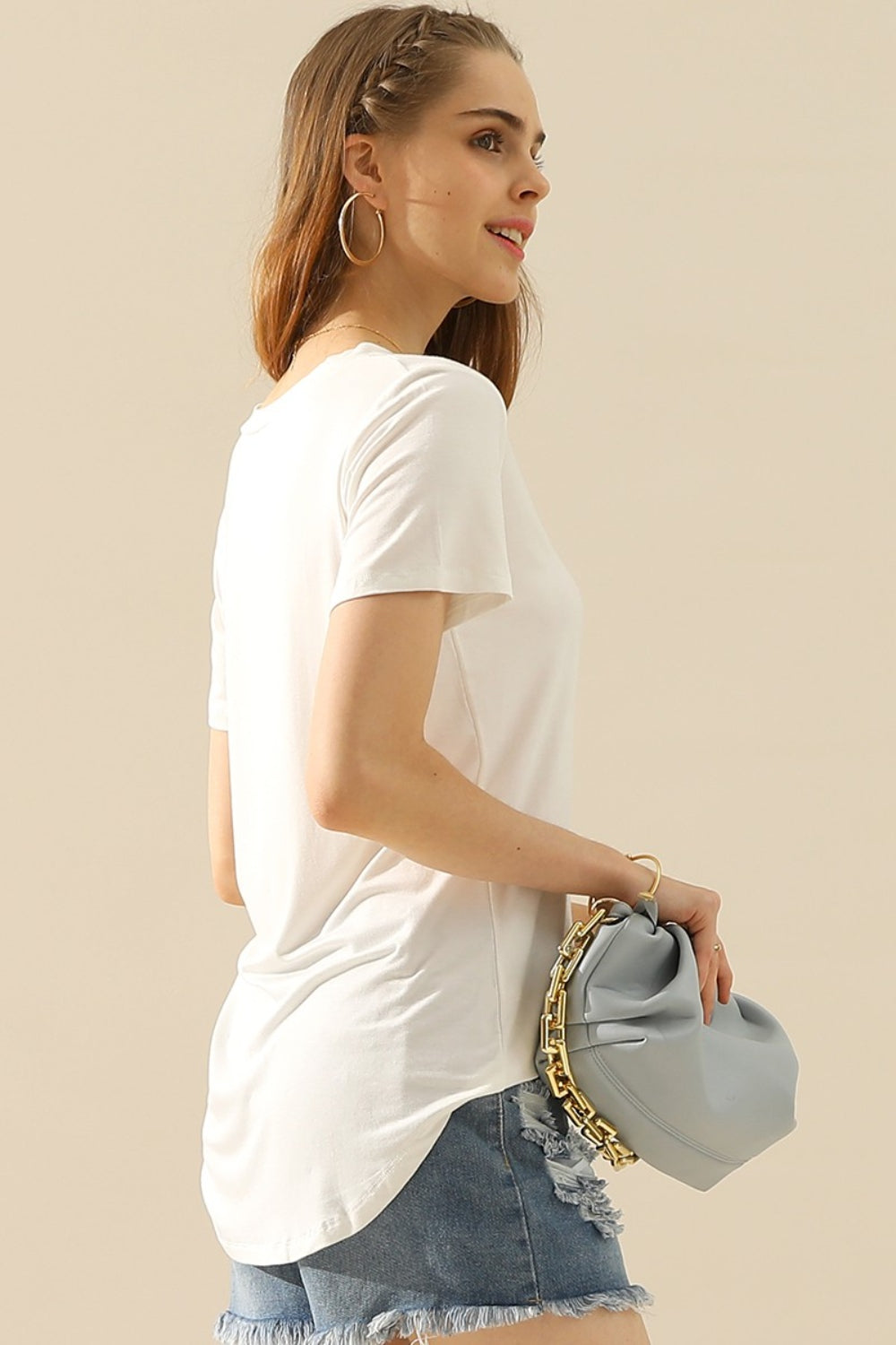 A woman with long hair wearing a Ninexis Full Size V-Neck Short Sleeve T-Shirt, a ripped denim skirt, and hoop earrings smiles at the camera.