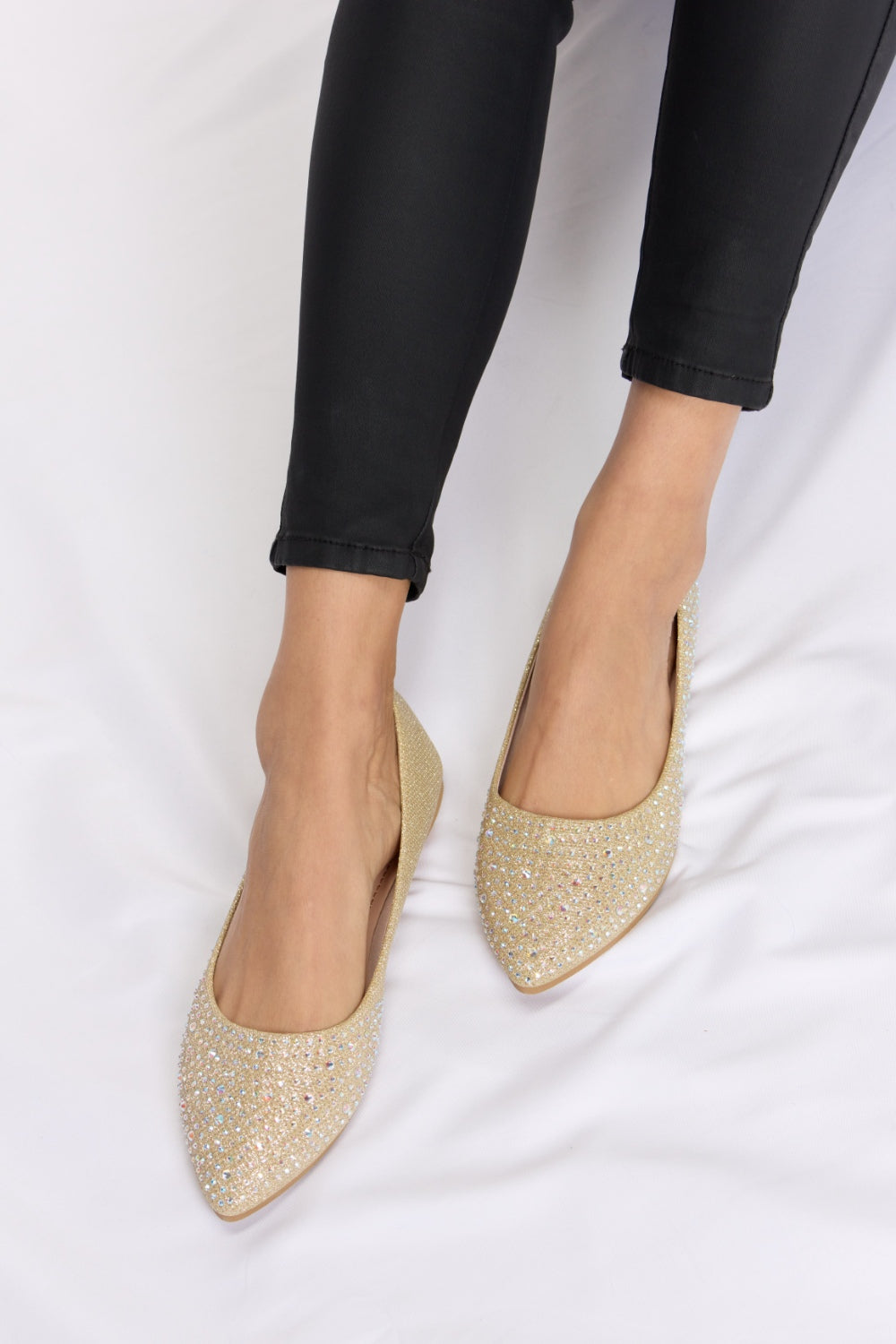 Someone is sitting on a white surface wearing black pants and chic Forever Link Rhinestone Point Toe Flat Slip-Ons in sparkling gold.