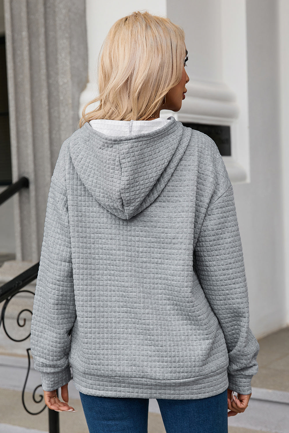 A person with long blonde hair is seen from behind, wearing a Gray Quilted Kangaroo Pocket Drawstring Hoodie and blue jeans. The relaxed fit of the textured fabric gives a casual vibe as they stand outdoors near a staircase with a metal railing.