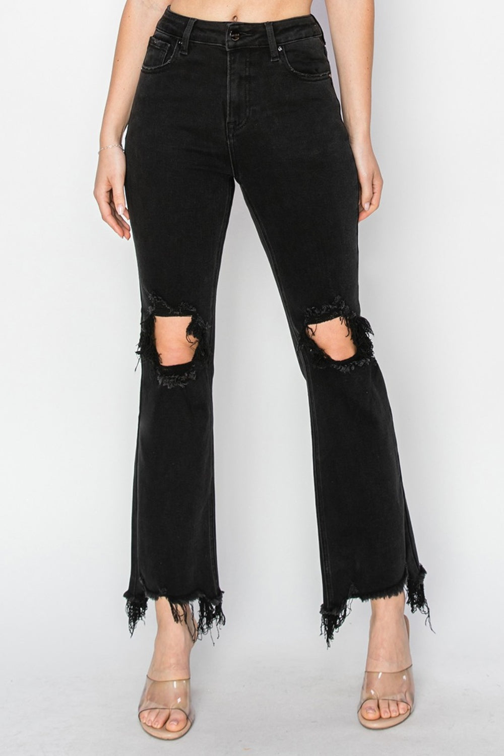 Rear view of a person wearing RISEN Full Size Distressed Raw Hem Jeans with Pockets in black, paired with clear-heeled sandals against a plain background.