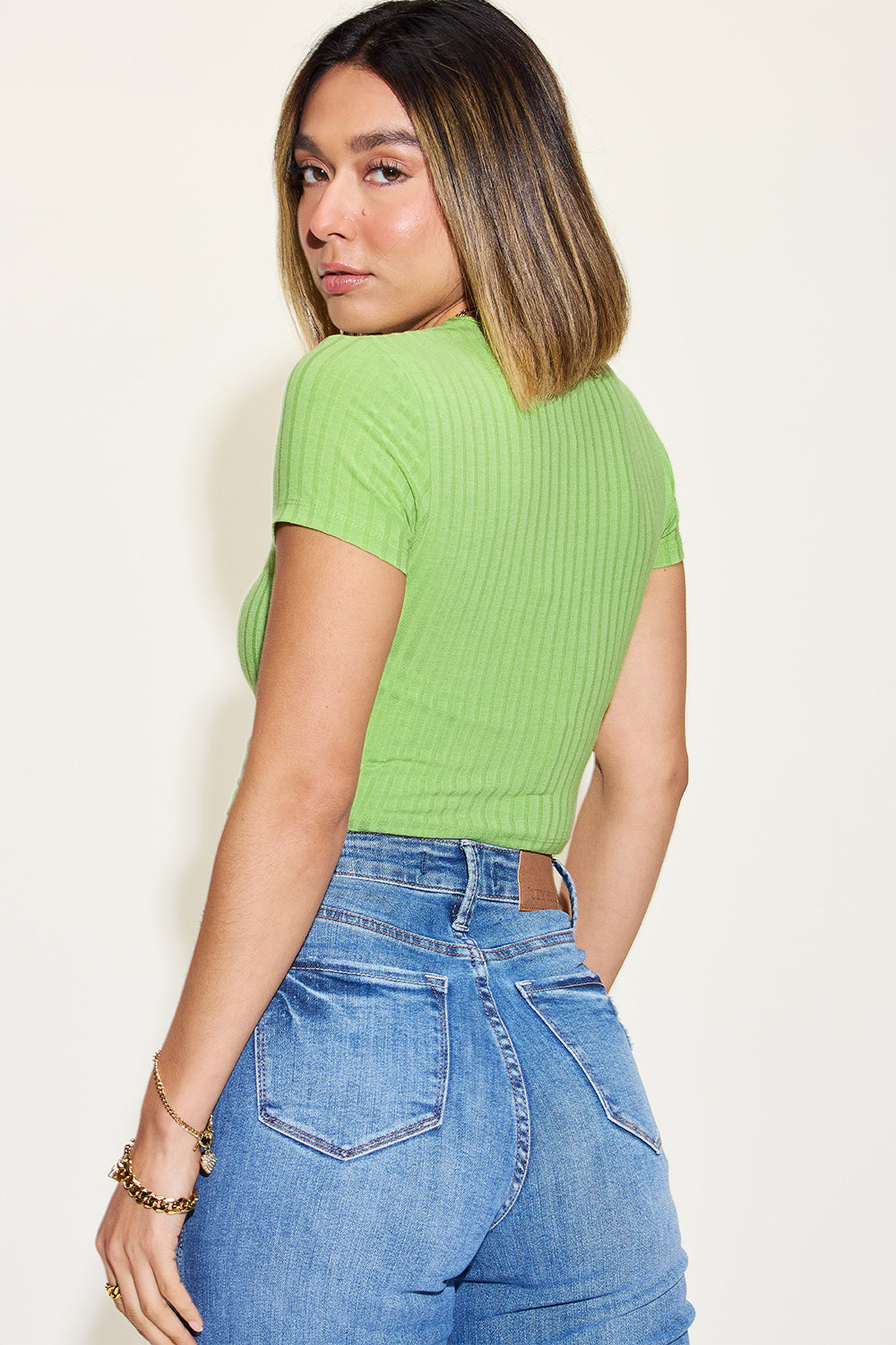 A woman with long blonde hair wearing a brown Basic Bae Full Size Ribbed Round Neck Short Sleeve T-Shirt, blue jeans, and gold jewelry stands against a plain light-colored background. The outfit boasts a basic style with moderate stretch for comfort.