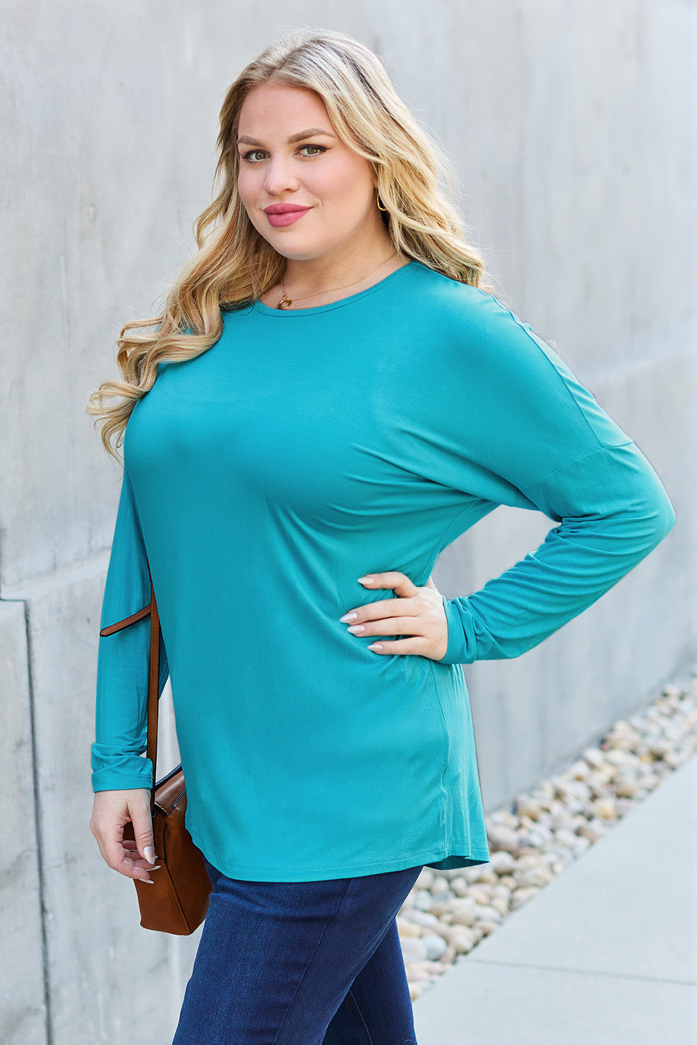 A woman with shoulder-length hair wearing a Basic Bae Full Size Round Neck Dropped Shoulder T-Shirt in green and a pair of jeans, exuding a basic style, stands against a concrete wall. She's holding an imported brown woven backpack on her shoulder while touching her hair with her hand.