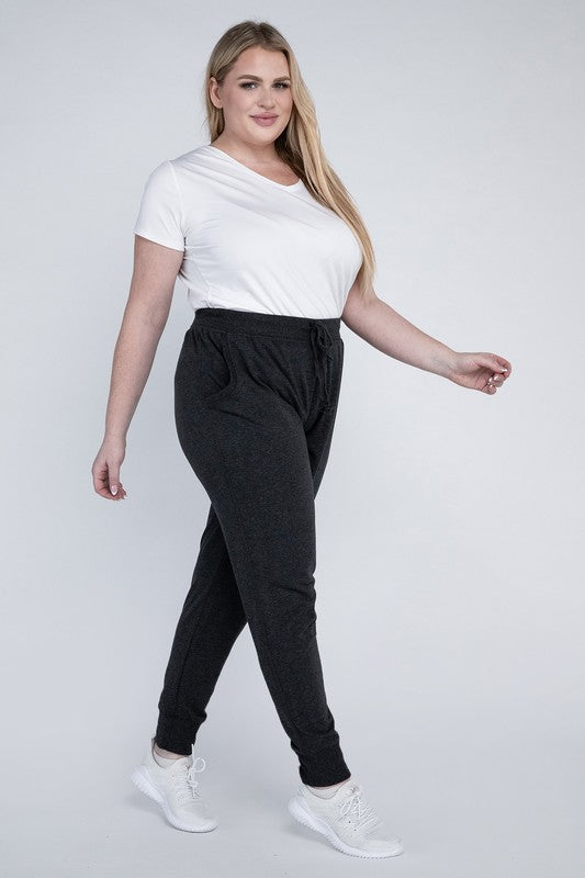 A person stands against a white background, wearing a white t-shirt, grey Plus-Size Jogger Pants with an adjustable waistband, and white sneakers. They have long blonde hair and are posing with one hand on their hip.