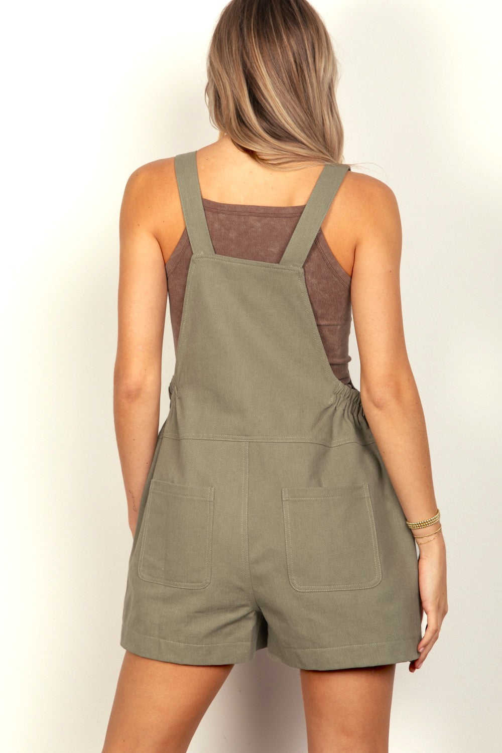 A woman with long blonde hair is wearing a pair of olive green VERY J Adjustable Suspender Overalls with an elastic waist, while her hands rest in the front pockets.