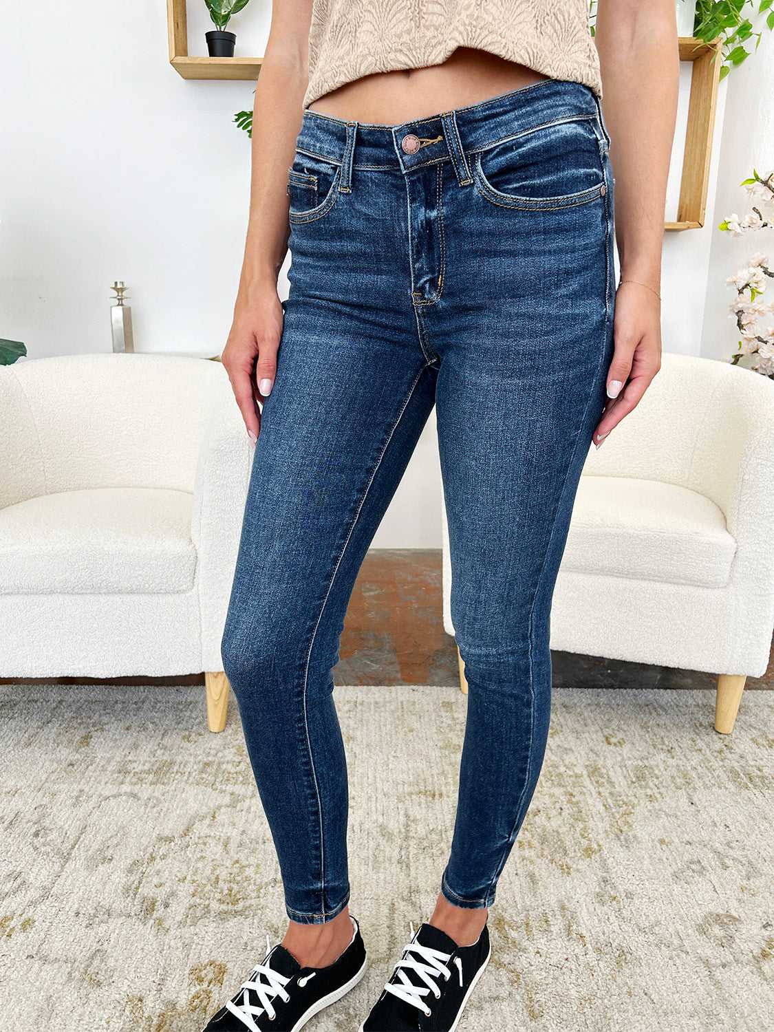 A person stands indoors wearing Judy Blue Full Size Classic Handsand Skinny Jeans, a beige top, and black sneakers with white laces. Their right hand is in their pocket. A partial view of two white chairs is visible in the background. The jeans feature a hand-sanded finish for a flattering fit.