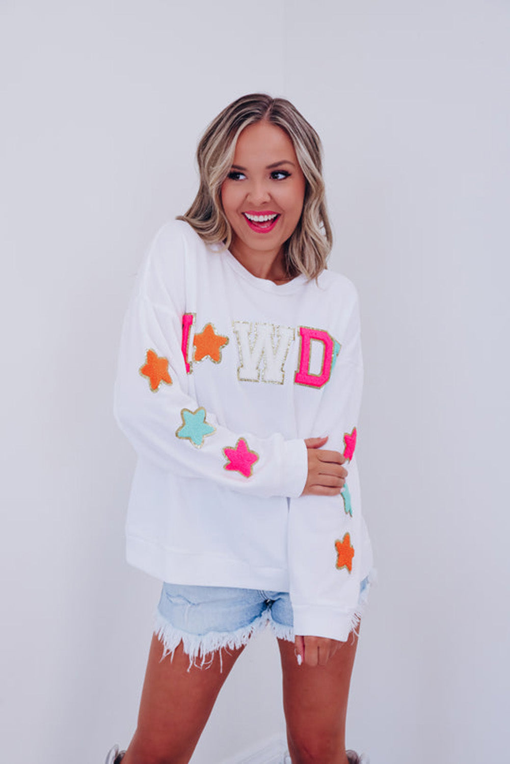 A person poses in the White Howdy Glitter Chenille Patch Graphic Casual Sweatshirt, adorned with colorful star patterns and the letters "WD." She wears light denim shorts and has shoulder-length blonde hair.